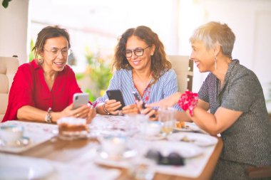 Orta yaşlı kadınlarla buluşup, öğle yemeği yiyip, kahve içiyorlar. Olgun arkadaşlar mutlu bir şekilde gülümsüyor. Güneşli bir günde evde akıllı telefon kullanıyorlar.