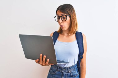 Beautiful redhead student woman wearing a backpack and using laptop scared in shock with a surprise face, afraid and excited with fear expression