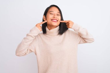 Young chinese woman wearing turtleneck sweater standing over isolated white background smiling cheerful showing and pointing with fingers teeth and mouth. Dental health concept.