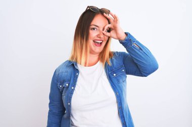 Güzel bir kadın kot gömlek giyiyor, beyaz arka plan üzerinde duruyor. El hareketleriyle gülümsüyor, gözleriyle mutlu yüzle parmaklarının arasından bakıyor..