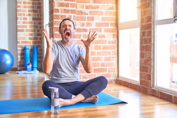 Middle age handsome sportman sitting on mat doing stretching yoga exercise at gym celebrating mad and crazy for success with arms raised and closed eyes screaming excited. Winner concept
