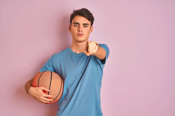 Teenager boy holding professional basket ball over isolated pink background pointing with finger to the camera and to you, hand sign, positive and confident gesture from the front