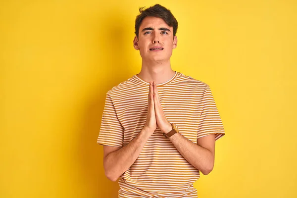 Teenager boy wearing yellow t-shirt over isolated background begging and praying with hands together with hope expression on face very emotional and worried. Asking for forgiveness. Religion concept.
