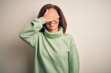 Orta yaşlı, güzel bir kadın, izole edilmiş beyaz arka plan üzerine balıkçı yaka kazak giyip, gülümseyerek ve yüzünde şaşkınlıktan gözleri kapayarak gülüyor. Kör kavram.