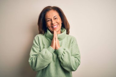 Orta yaşlı, güzel, balıkçı yaka kazak giyen, soyutlanmış beyaz arka planda dua eden ellerini birleştirip af dileyen, kendinden emin bir şekilde gülümseyen bir kadın..