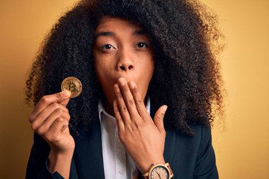 Afro saçlı Afrikalı Amerikalı genç bir iş kadını sarı arka planda Bitcoin tutuyor. Ağzı kapalı, utancından ağzı açık, korkunun ifadesi, sessizlikten korkan, gizli bir konsept.