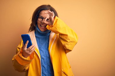 Sarı yağmurluk giyen orta yaşlı bir kadın izole edilmiş sarı arka plan üzerinde akıllı telefon kullanıyor. Gülümsüyor. Tamam işaret veriyor.