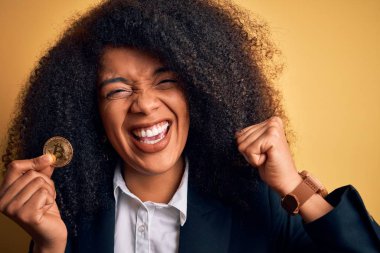 Afro saçlı Afrikalı Amerikalı genç bir iş kadını sarı arka planda bitcoin tutuyor gururla bağırıyor ve zaferi ve başarıyı kutluyor çok heyecanlı, coşkulu bir duygu.