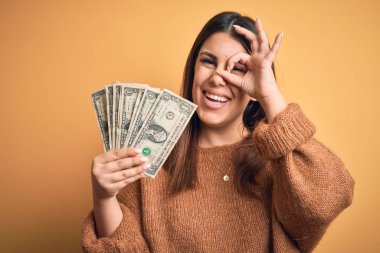 Dolarları tutan genç ve güzel bir kadın mutlu yüzüyle turuncu arka planda duruyor. Elleri gözlerinde parmaklarının arasından bakıyor.