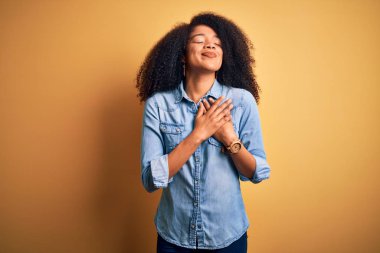 Sarıya boyanmış arka planda duran, elleri göğsünde, gözleri kapalı ve yüzünde minnet dolu bir jestle gülümseyen Afro saçlı güzel Afrikalı Amerikalı kadın. Sağlık kavramı.