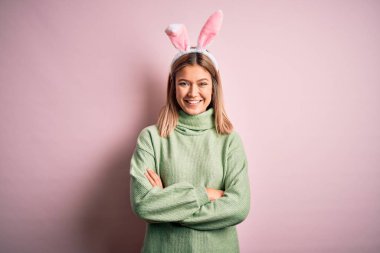 Paskalya tavşanı kulakları takan genç ve güzel bir kadın izole edilmiş pembe arka plan üzerinde durup kollarını kavuşturup kameraya bakarak gülümsüyor. Pozitif kişi.
