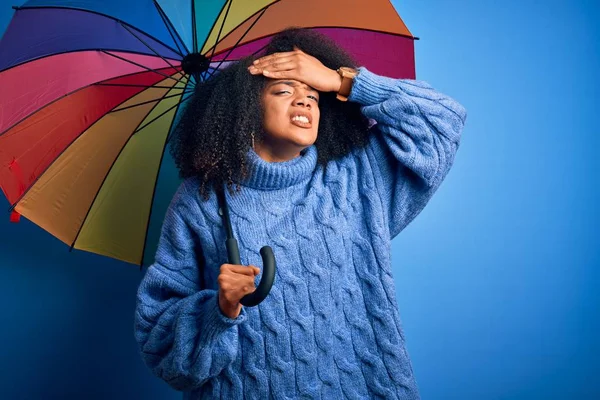 Young african american woman with afro hair under colorful umbrella for winter weather rain stressed with hand on head, shocked with shame and surprise face, angry and frustrated. Fear and upset for mistake.