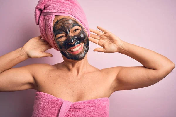 Middle age brunette woman wearing beauty black face mask over isolated pink background relaxing and stretching, arms and hands behind head and neck smiling happy