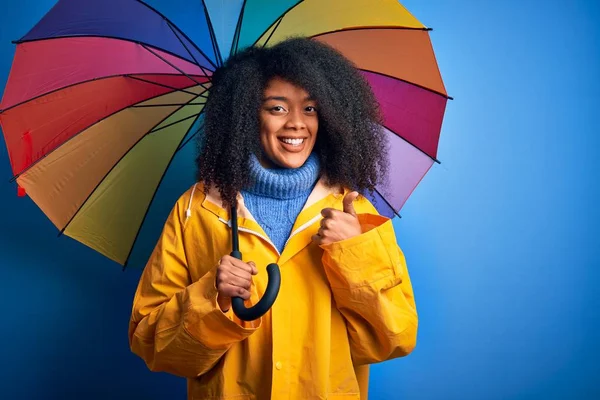 Young african american woman with afro hair under colorful umbrella wearing winter coat for rain happy with big smile doing ok sign, thumb up with fingers, excellent sign