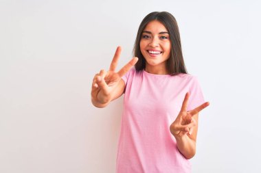 İzole edilmiş beyaz arka planda duran pembe günlük tişört giyen genç ve güzel bir kadın kameraya bakıp zafer işareti yapıyor. İki numara..