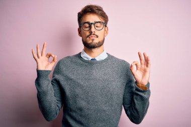 Sakallı, gözlüklü ve süveterli genç bir adam pembe arka planda dikiliyor rahatlıyor ve parmaklarıyla meditasyon yaparken gözleri kapalı gülümsüyor. Yoga konsepti.