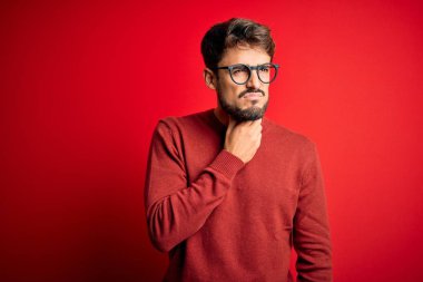 Sakallı, gözlüklü ve süveterli genç adam kırmızı arka planda duruyor. Ağrılı boynuna dokunuyor. Grip, mantar ve enfeksiyon yüzünden boğazı ağrıyor.