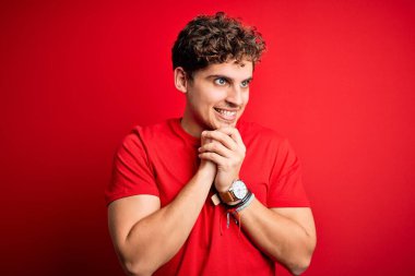 Sarışın, kıvırcık saçlı, kırmızı arka planda sıradan tişörtler giyen, elleri çene hizasında gergin ve heyecanlı genç bir adam.