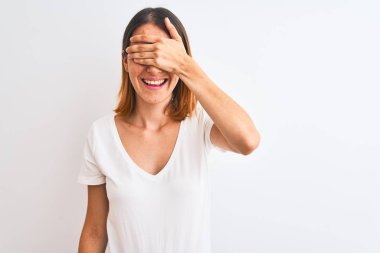 Güzel kızıl saçlı kadın, izole edilmiş arka planda günlük beyaz tişörtler giyip gülümsüyor ve elleri yüzünde şaşkınlıktan gözleri kapayarak gülüyor. Kör kavram.