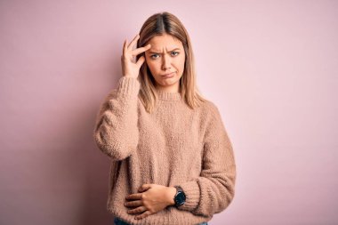 Pembe izole edilmiş arka planda, kışlık yün kazak giyen genç, güzel sarışın kadın. Alnındaki sorun yüzünden endişeli ve stresli. Kriz konusunda endişeli.