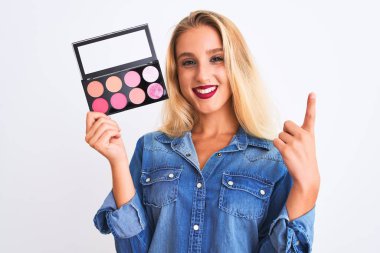 Makyaj yapan genç, güzel, sarışın bir kadın izole edilmiş beyaz arka planda duruyor. Bir fikir ya da soru, mutlu bir yüzle işaret ediyor.