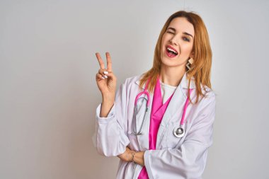 İzole edilmiş arka planda pembe steteskop takan Kafkasyalı doktor kadın kameraya bakıp gülümsüyor ve zafer işareti yapıyor. İki numara..