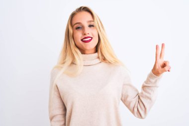 Balıkçı yaka kazak giyen genç ve güzel bir kadın izole edilmiş beyaz arka plan üzerinde duruyor ve parmaklarını iki numara ile gösterirken kendinden emin ve mutlu bir şekilde gülümsüyor..