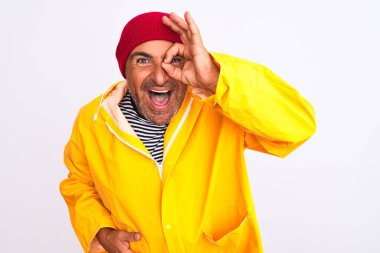 Middle age man wearing rain coat and woolen hat standing over isolated white background doing ok gesture with hand smiling, eye looking through fingers with happy face.