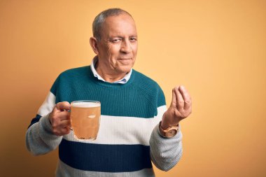 Senior handsome man drinking jar of beer standing over isolated yellow background doing money gesture with hands, asking for salary payment, millionaire business