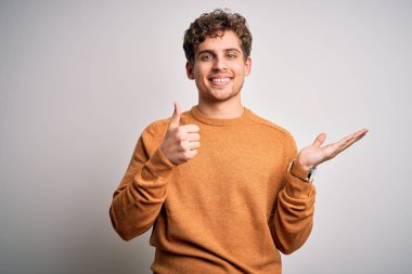 Sarışın, kıvırcık saçlı, beyaz arka planda sıradan bir süveter giyen, avucunun içini gösteren ve baş parmaklarıyla hareket eden mutlu ve neşeli gülümseyen genç bir adam.