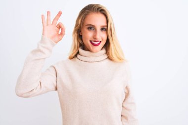 Young beautiful woman wearing turtleneck sweater standing over isolated white background smiling positive doing ok sign with hand and fingers. Successful expression.