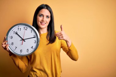 Young woman with blue eyes doing countdown holding big clock over yellow background happy with big smile doing ok sign, thumb up with fingers, excellent sign