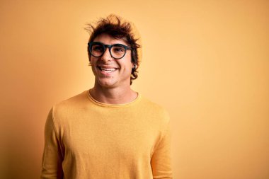 Young handsome man wearing casual t-shirt and glasses over isolated yellow background looking away to side with smile on face, natural expression. Laughing confident.