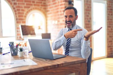 Middle age handsome businessman wearing tie sitting using laptop at the office amazed and smiling to the camera while presenting with hand and pointing with finger.