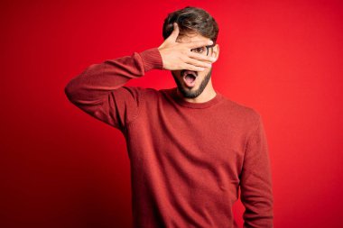 Young handsome man with beard wearing glasses and sweater standing over red background peeking in shock covering face and eyes with hand, looking through fingers with embarrassed expression.