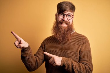 Handsome Irish redhead man with beard wearing glasses and winter sweater over yellow background smiling and looking at the camera pointing with two hands and fingers to the side.