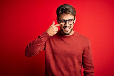 Young handsome man with beard wearing glasses and sweater standing over red background Pointing with hand finger to face and nose, smiling cheerful. Beauty concept
