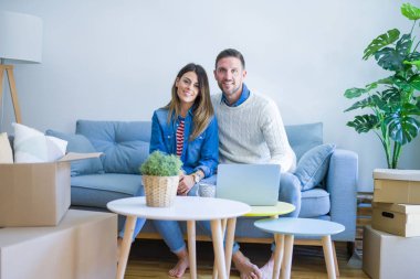 Genç güzel çift karton kutular etrafında yeni evde dizüstü bilgisayar kullanarak kahve kanepede içme fincan oturan