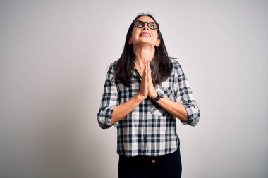 Young brunette woman with blue eyes wearing casual shirt and glasses over white background begging and praying with hands together with hope expression on face very emotional and worried. Begging.