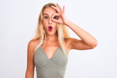 Young beautiful woman wearing casual green t-shirt standing over isolated white background doing ok gesture shocked with surprised face, eye looking through fingers. Unbelieving expression.
