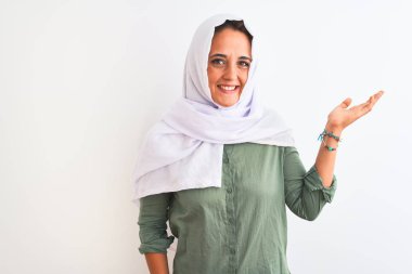 Young beautiful Arab woman wearing traditional Muslim hijab over isolated background smiling cheerful presenting and pointing with palm of hand looking at the camera.