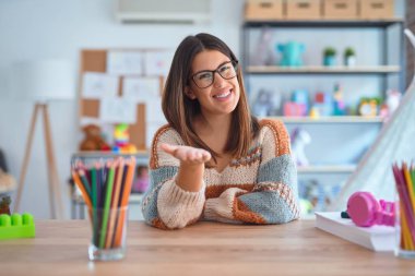 Young beautiful teacher woman wearing sweater and glasses sitting on desk at kindergarten smiling cheerful offering palm hand giving assistance and acceptance. clipart