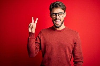 Young handsome man with beard wearing glasses and sweater standing over red background smiling with happy face winking at the camera doing victory sign with fingers. Number two.