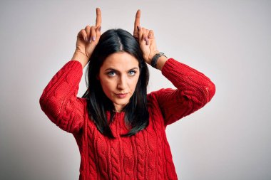 Young brunette woman with blue eyes wearing casual sweater over isolated white background doing funny gesture with finger over head as bull horns