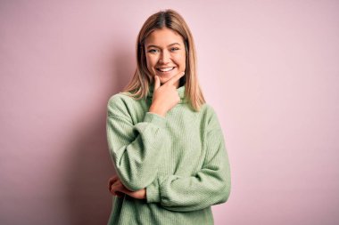 Young beautiful blonde woman wearing winter wool sweater over pink isolated background looking confident at the camera smiling with crossed arms and hand raised on chin. Thinking positive.