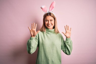 Young beautiful woman wearing easter rabbit ears standing over isolated pink background showing and pointing up with fingers number ten while smiling confident and happy.