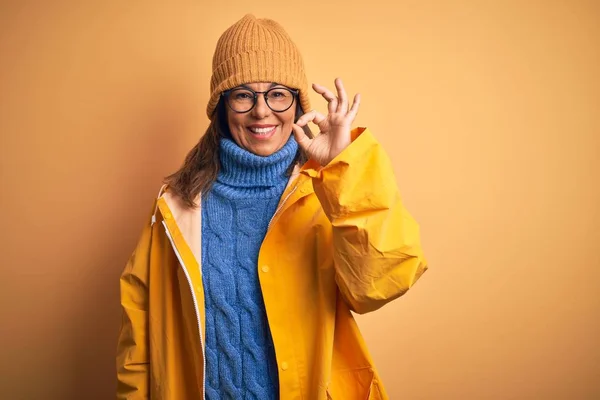 Middle age woman wearing yellow raincoat and winter hat over isolated background smiling positive doing ok sign with hand and fingers. Successful expression.