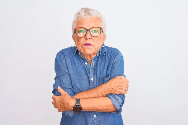 Senior grey-haired woman wearing denim shirt and glasses over isolated white background shaking and freezing for winter cold with sad and shock expression on face