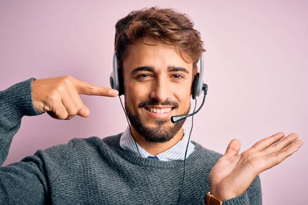 Young call center agent man with beard wearing headset over isolated pink background amazed and smiling to the camera while presenting with hand and pointing with finger.