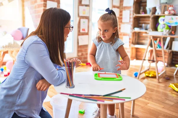 Beyaz kız çocuk, bayan öğretmenle oyun oynayıp öğreniyor. Anne ve kızı oyun odasında oyuncakların etrafında manyetik karatahtaya resim çiziyorlar.